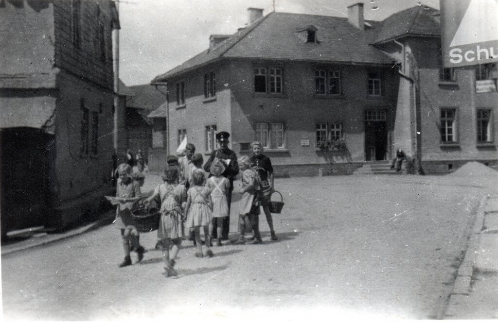 Postbote Julius Fischer mit Schulkindern auf der Hauptstraße (später Schwalbacher Straße). Im Hintergrund die Schule, links das Anwesen Leichtfuß / Rechener. Foto von Irmgard Pfeil.